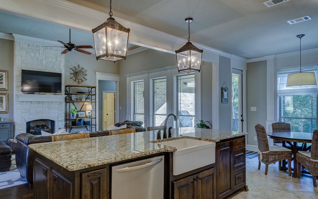 Open-plan kitchen with a granite island and farmhouse sink beside a stone fireplace and dining area.