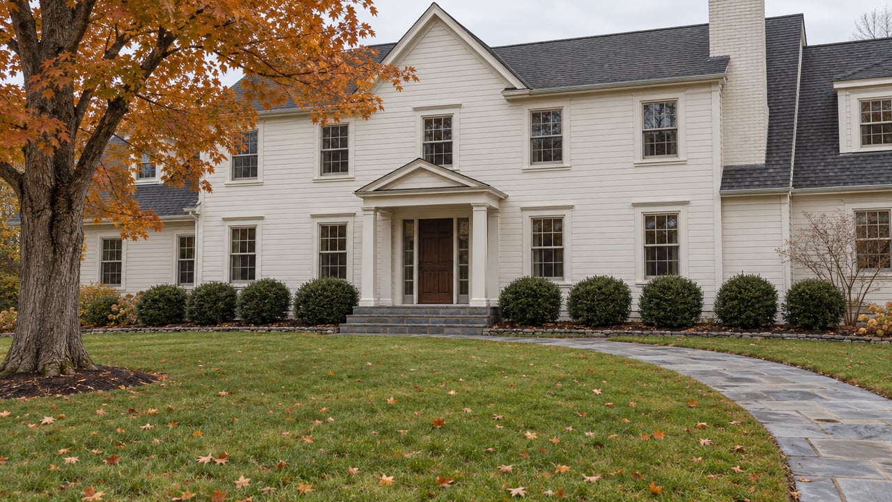 Light clapboard home with dark gabled roofs, a bluestone approach and a broad autumn lawn beside an orange-leaved maple.