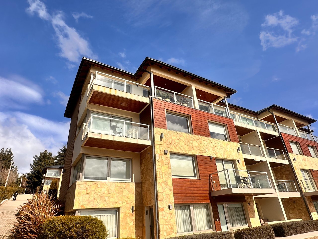 Apartment building with stone and wood siding, glass balconies, and outdoor seating beneath a blue sky.