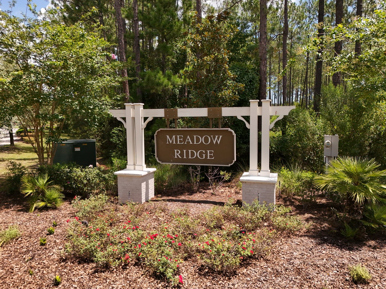 Meadow Ridge entrance sign surrounded by flowering shrubs and tall pine trees.