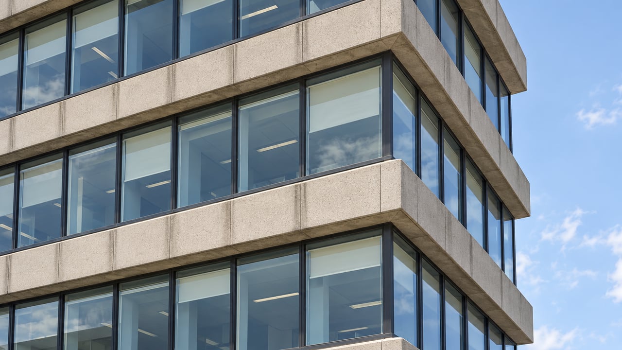 Low-angle close view of pale concrete office floors, charcoal window frames and blue-green glazing against a narrow strip of sky.