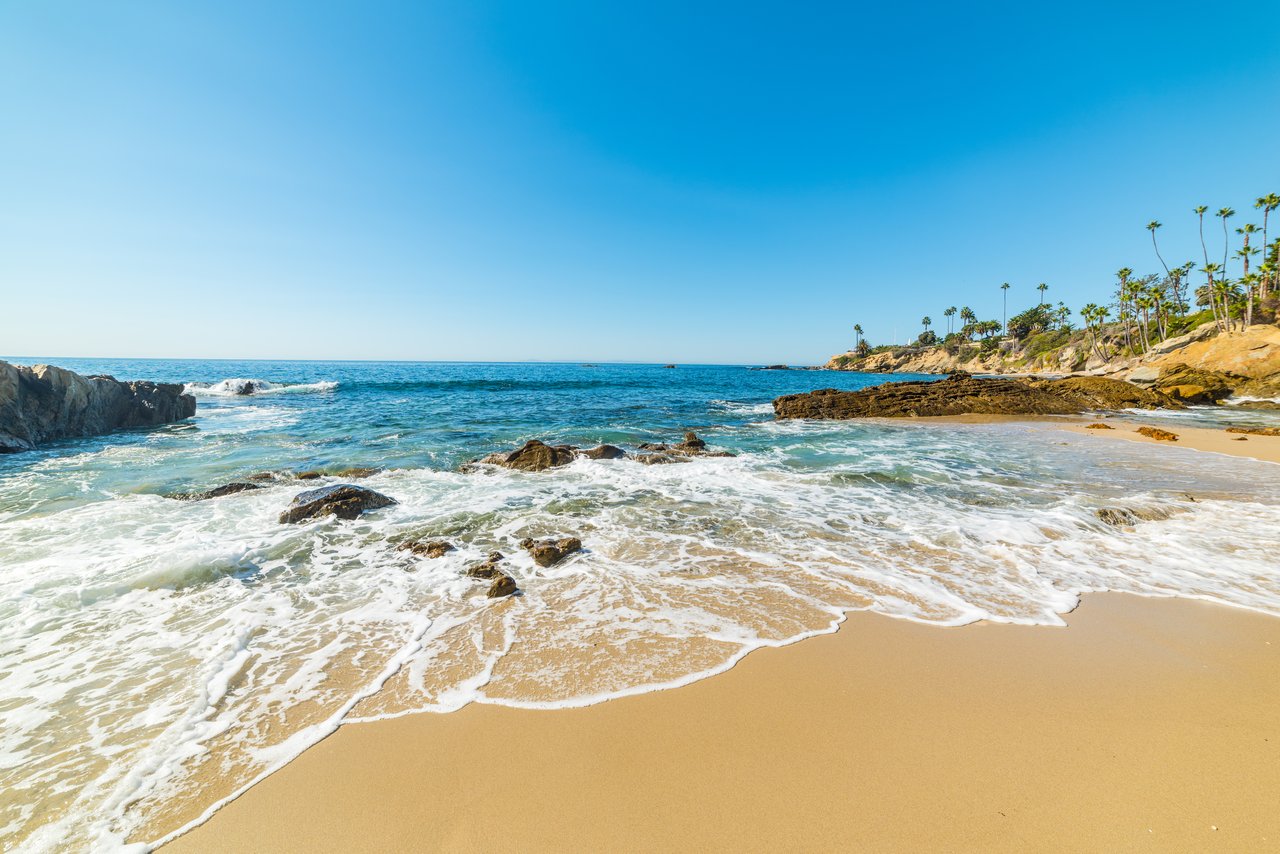 Sandy beach with waves breaking around rocks below a palm-lined headland.