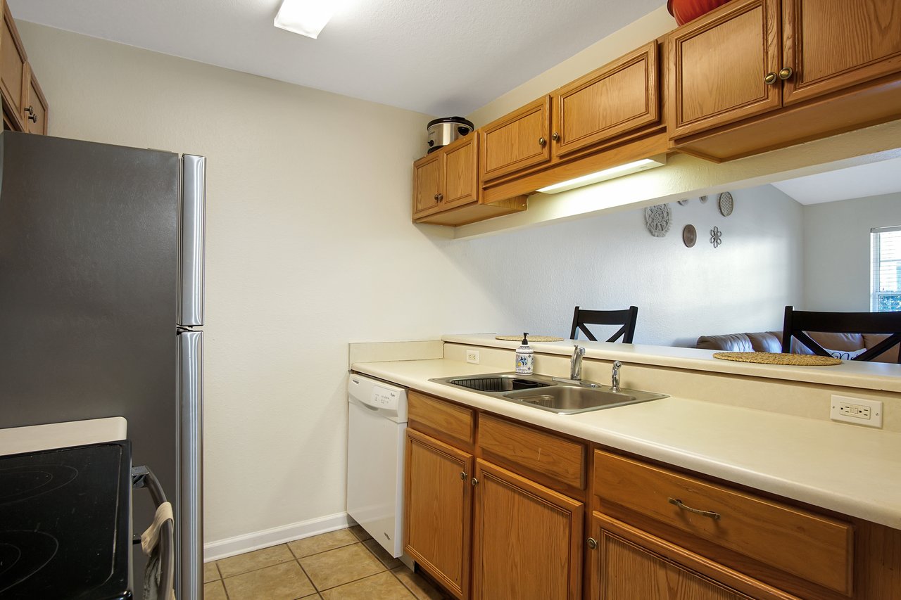 Compact kitchen with a stainless steel fridge, light wood cabinets, and a sink opposite bar stools. Tiled floor, beige walls, clean, and inviting.