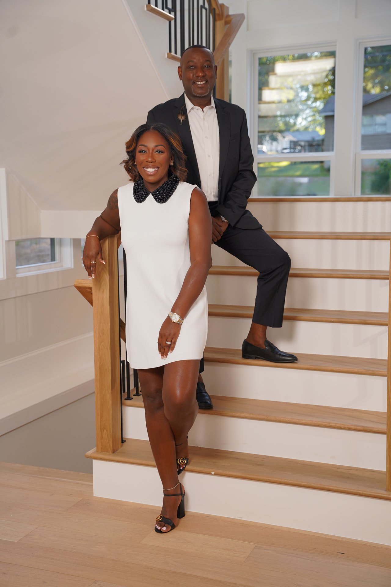 Two real estate professionals pose on a wooden staircase inside a bright home.