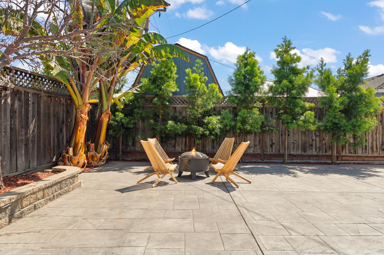 Backyard patio with wooden lounge chairs around a fire pit, framed by banana trees and a privacy fence.