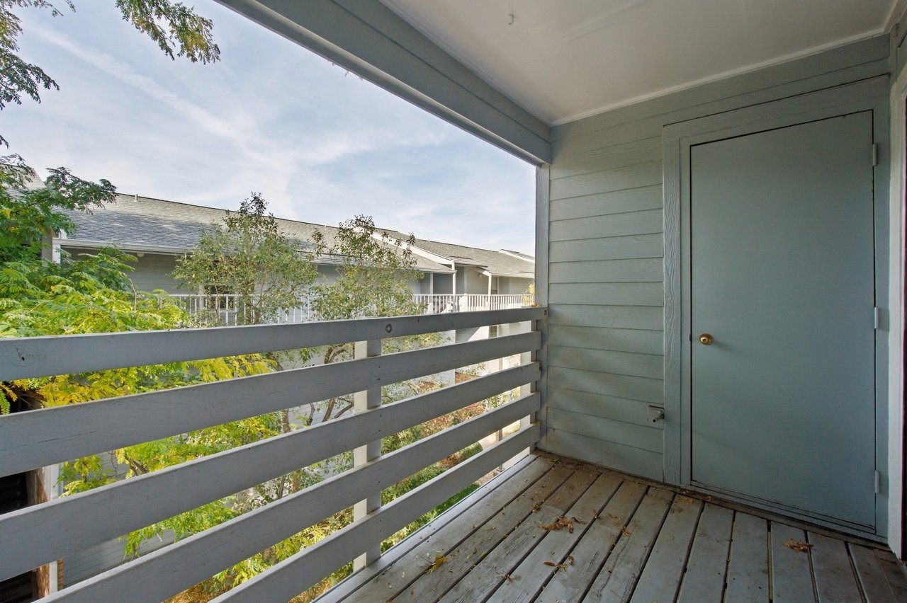 Covered balcony with gray railings and plank flooring overlooking trees and a neighboring building.