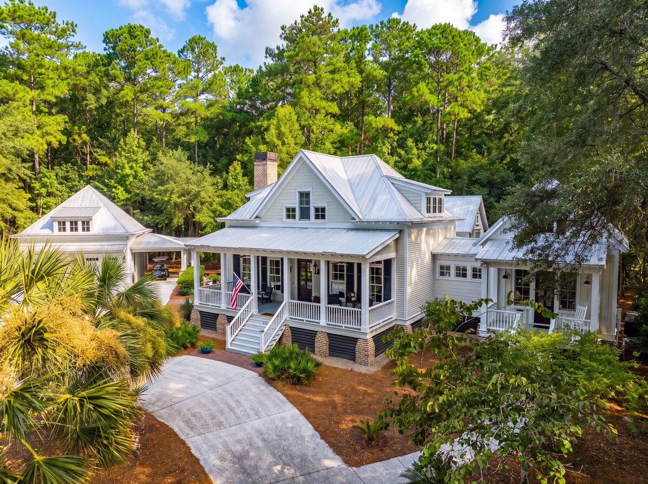 White cottage with a metal roof, broad porches, and a curved driveway among trees.