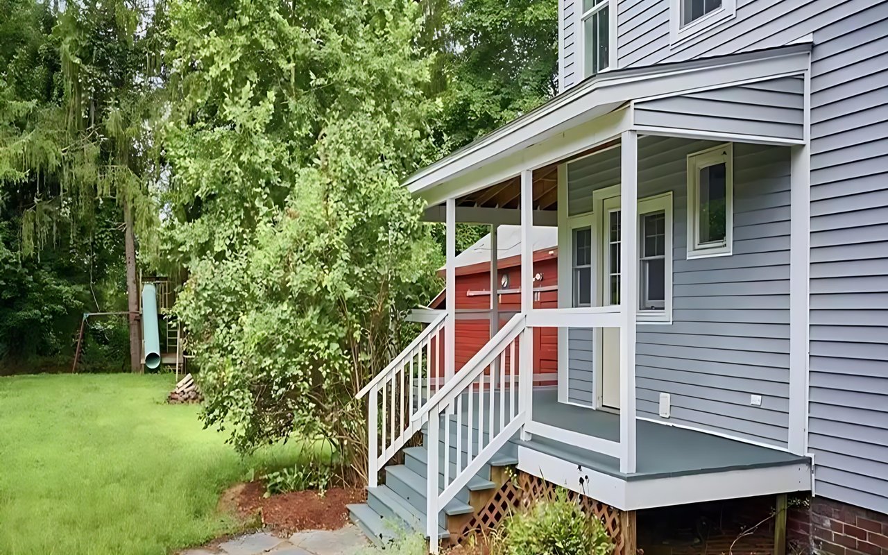 Gray-sided house with a covered porch and white railings beside a leafy yard.
