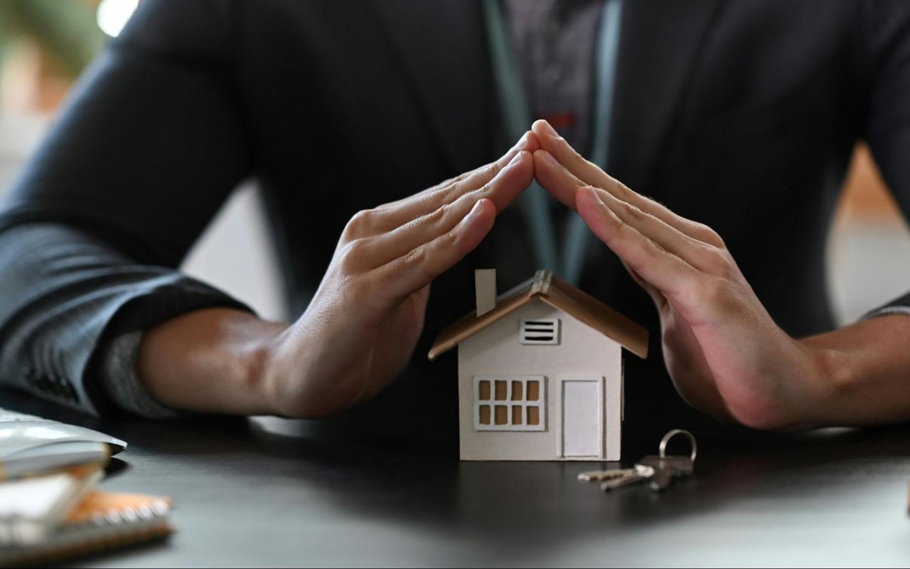  A person in a dark suit is holding their hands in a roof-like shape over a small, miniature house model. 