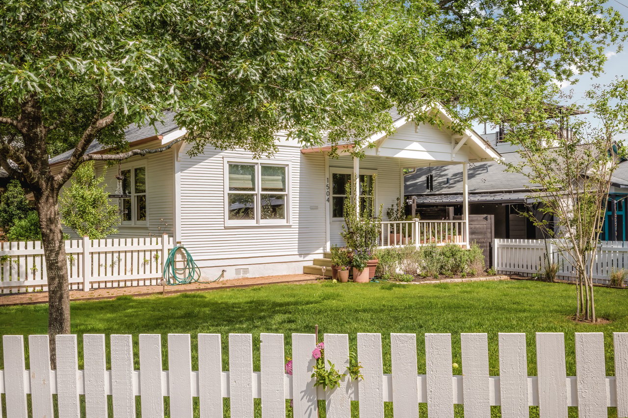 White single-story house behind a picket fence, shaded by a large leafy tree.