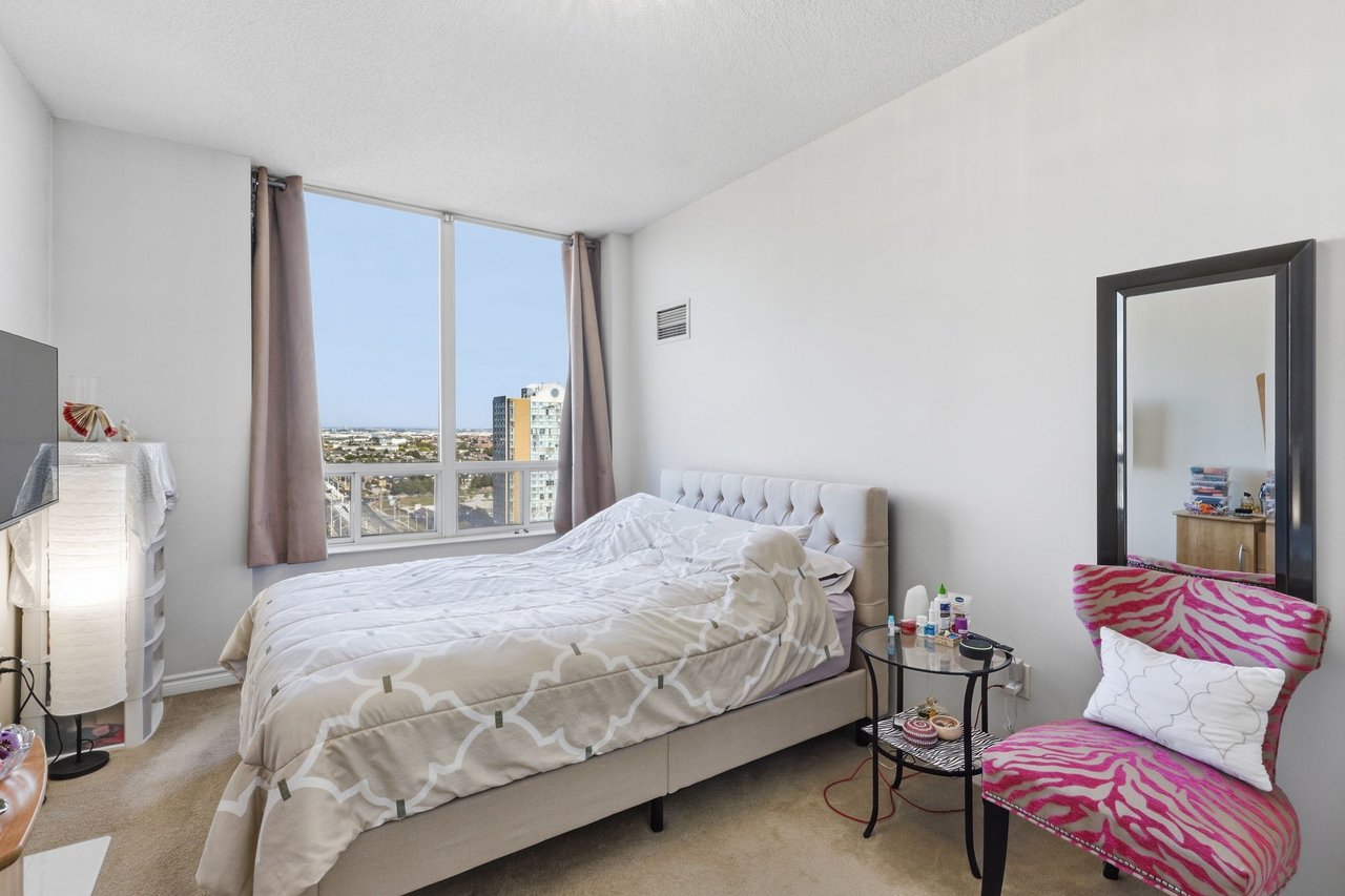 Furnished bedroom with a large bed, floor-to-ceiling city-view windows, and a bright pink patterned chair.