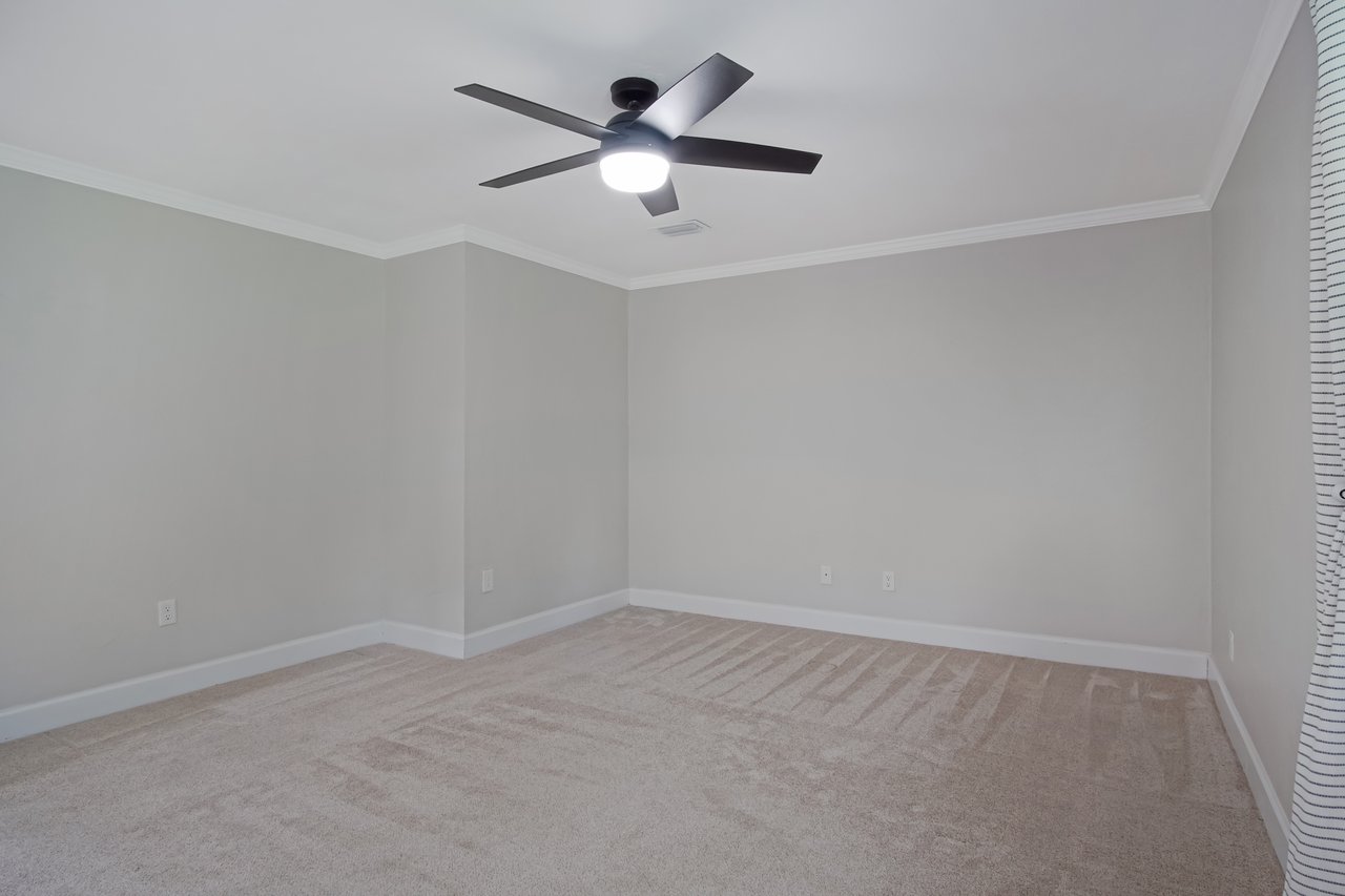 Empty gray room with carpeted floor, white trim, and a ceiling fan with light. A window with striped curtains is partially visible. Calm and minimalistic.