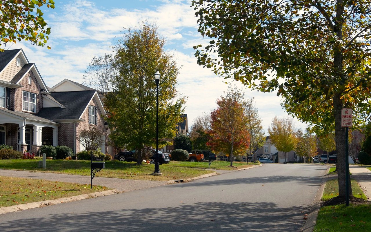 Residential street lined with homes and autumn trees, with a brick house in the foreground.