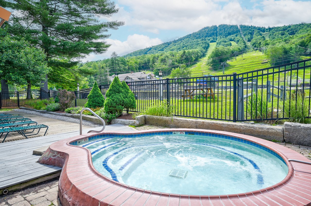 Outdoor hot tub beside a pool, with a forested mountain ski slope beyond the fence.