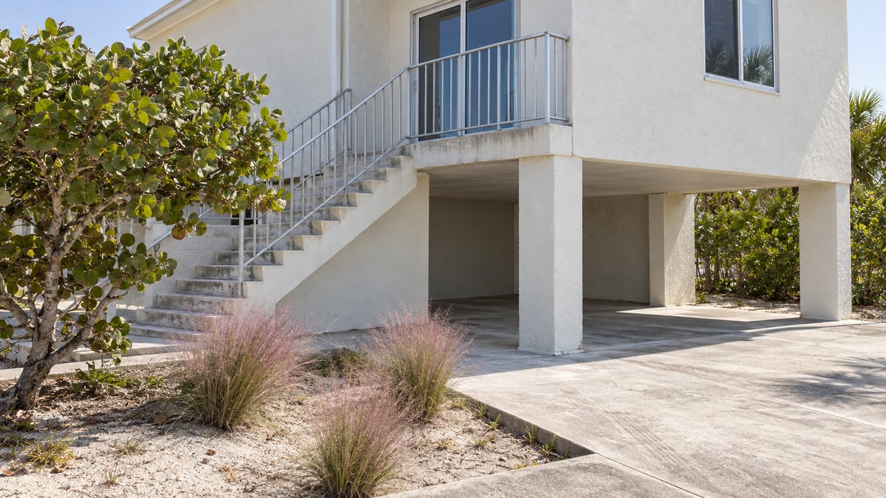 Raised coastal house with open parking beneath, concrete entry steps, and a sandy bed with grasses.