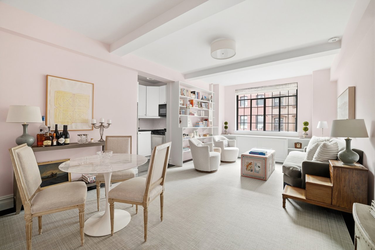 Open-plan living and dining room with pale pink walls, white lounge chairs, and a large city-facing window.