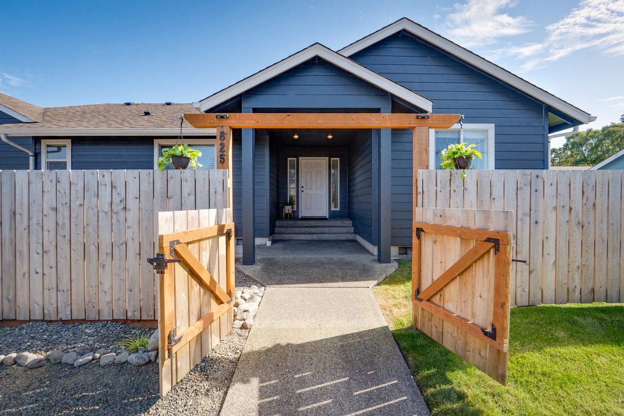 Front entrance of a modern single-story home in Seaside, Oregon, featuring dark blue siding, a covered entryway with wood accents, an open wooden gate, privacy fencing, and a concrete walkway leading to the front door.
