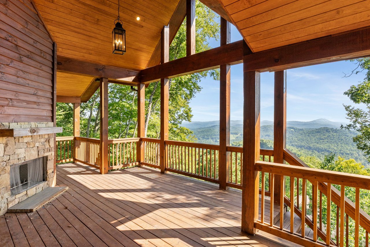 Covered wraparound deck at 523 Broadview with a tongue-and-groove wood ceiling, timber-frame posts and beams, a hanging lantern, a stacked-stone outdoor fireplace, and long-range Blue Ridge mountain views over the treetops.
