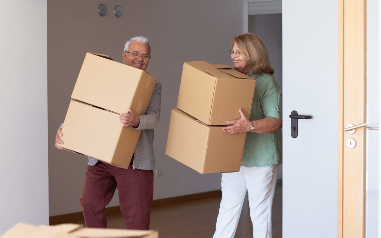 Older couple carrying stacked moving boxes through a doorway