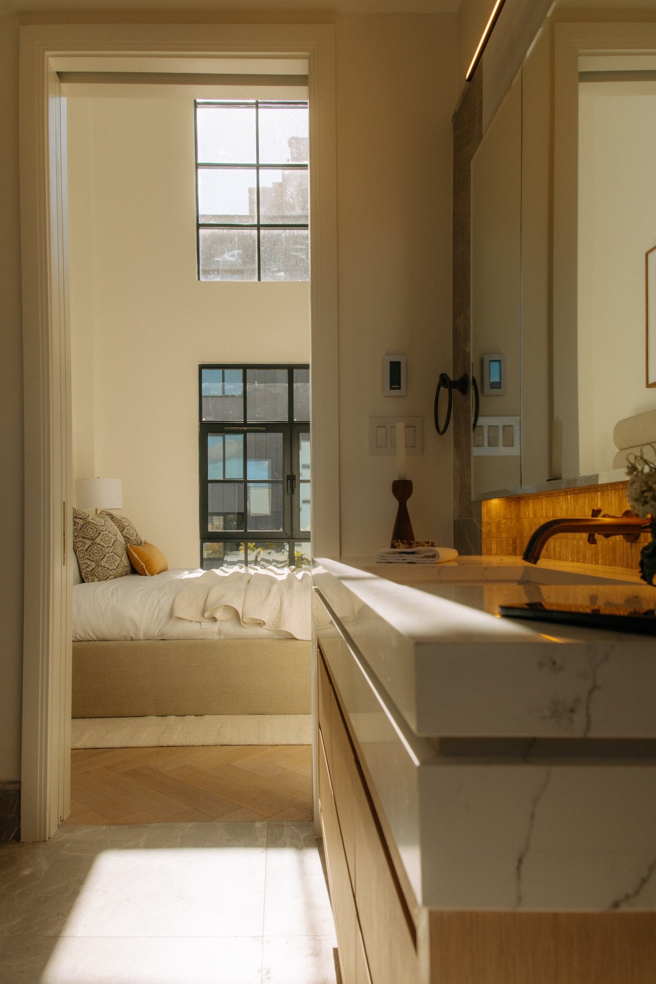 Sunlit bathroom with a marble vanity overlooking a bedroom with tall black-framed windows.