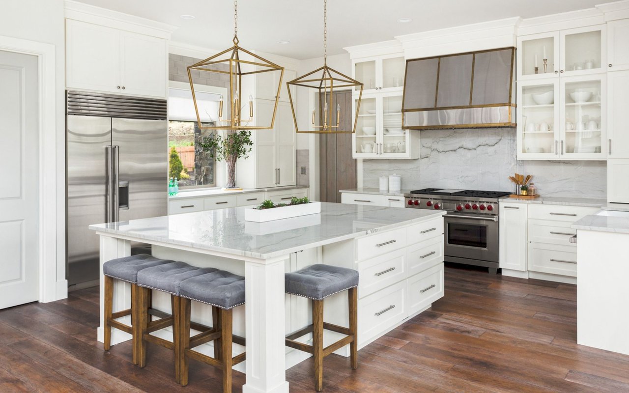 Bright white kitchen with a large marble island, gray bar stools, and stainless steel appliances.