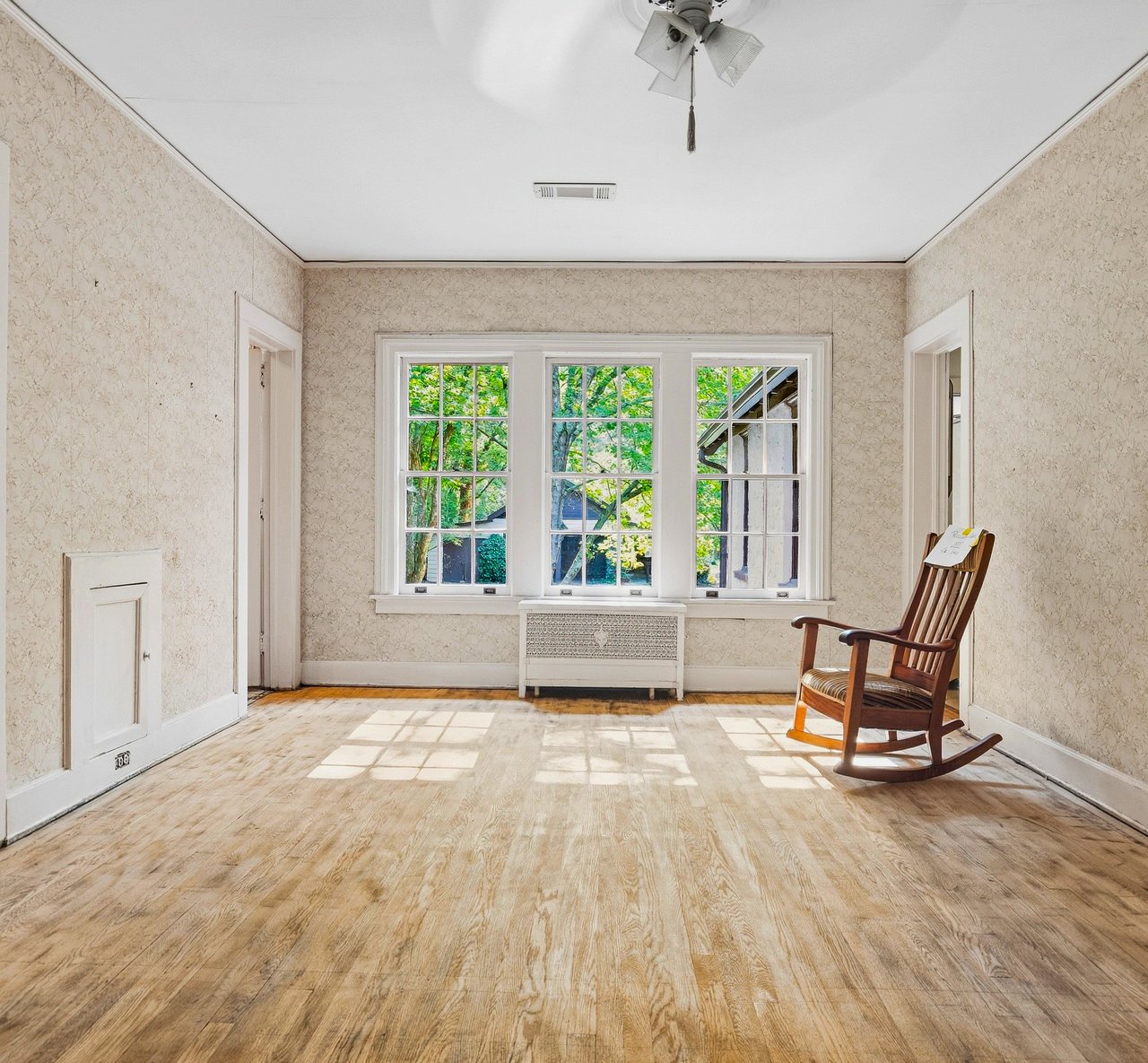 Mostly empty room with patterned wallpaper, wood floors, three large windows, and a wooden rocking chair.