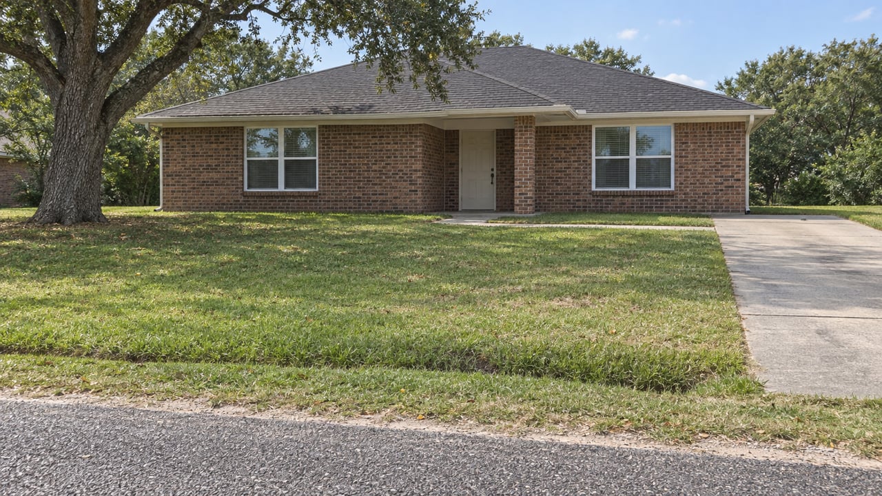 A modest brick ranch behind a deep lawn and shallow roadside swale, with a concrete driveway and mature live oak.