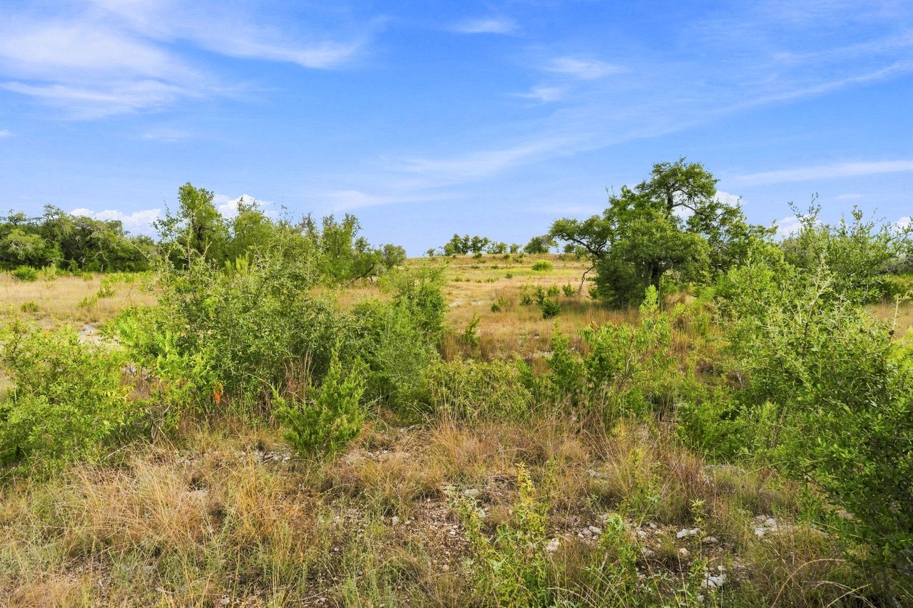 Open ranch acreage with rocky grassland, scattered shrubs, and mature oak trees beneath a blue sky.