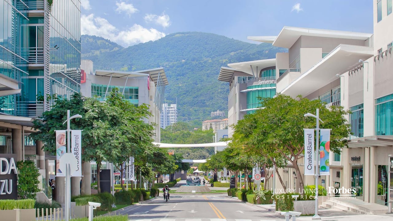 Avenida Escazú’s modern commercial buildings and tree-lined street, with green mountains in the distance.