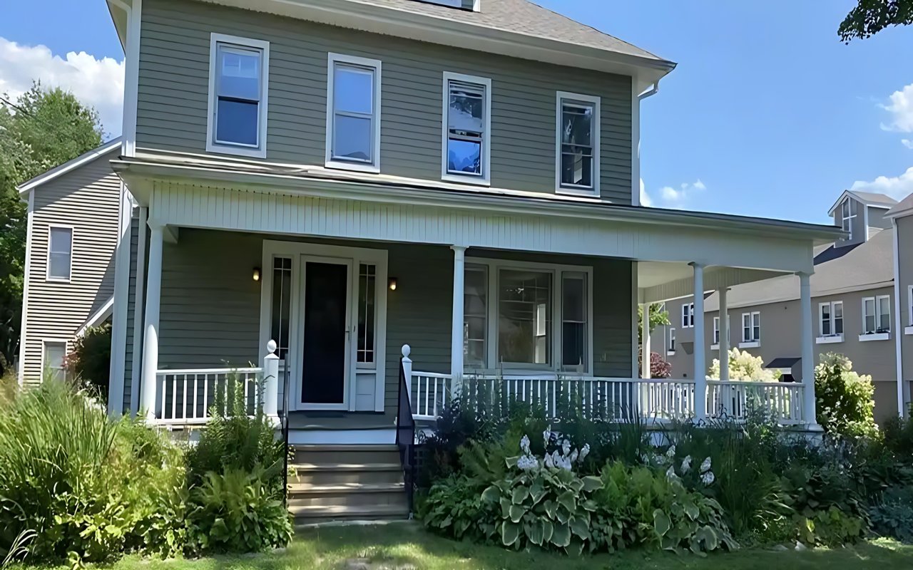 Two-story gray-green house with a broad covered porch and landscaped front garden.