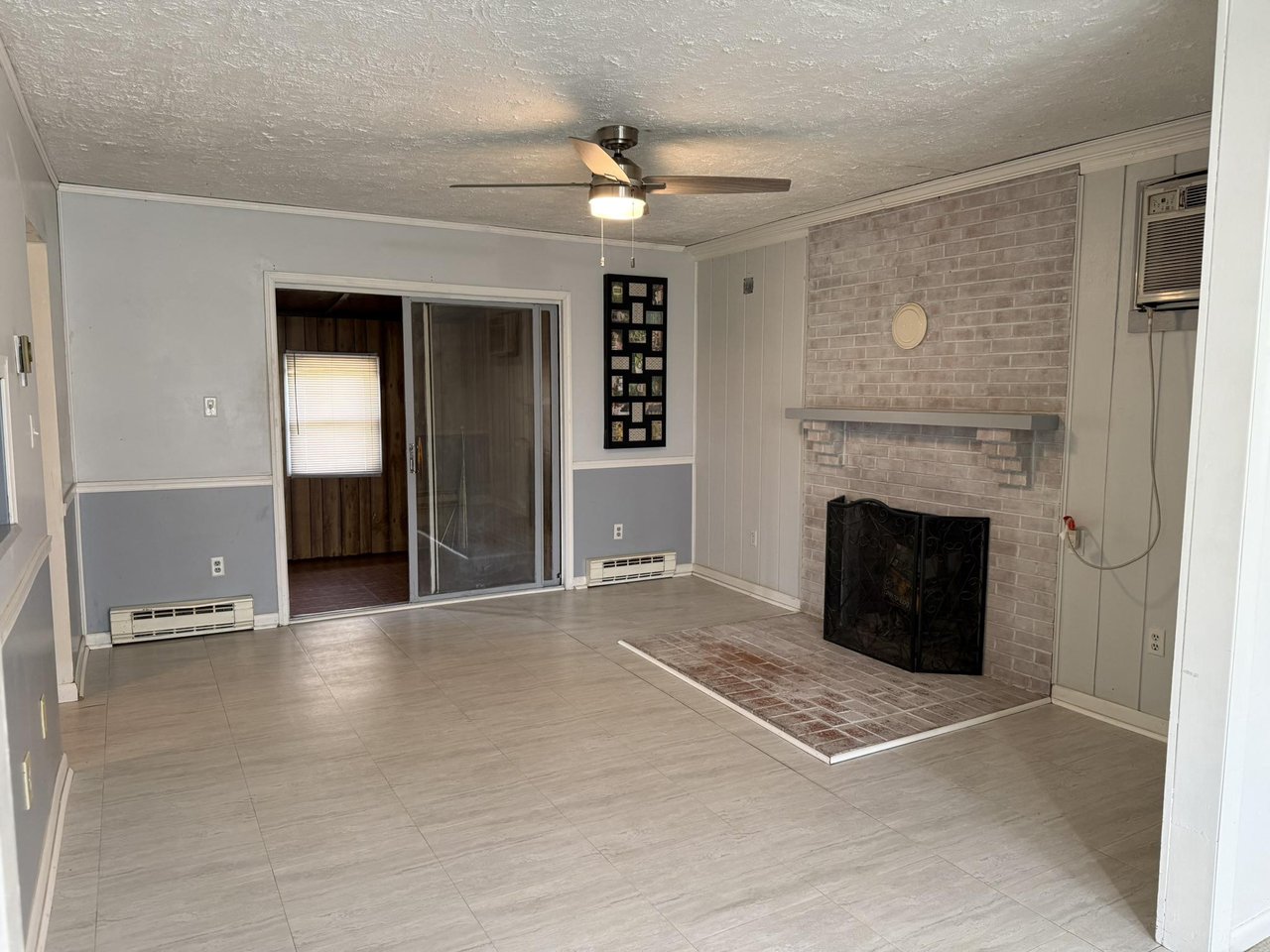 Empty living room with a pale brick fireplace, tiled floor, ceiling fan, and sliding glass doors.
