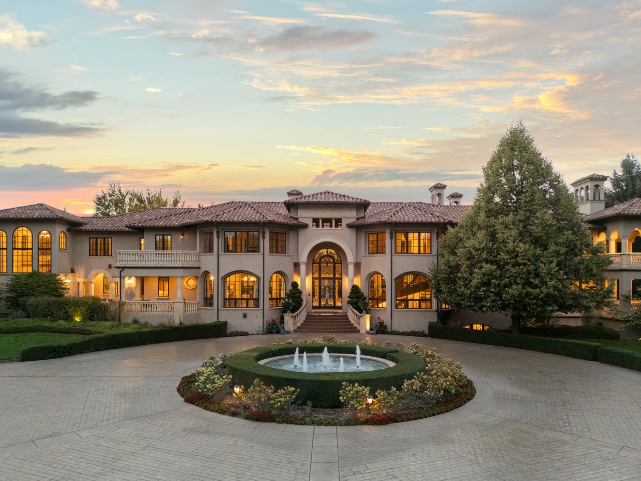 Mediterranean-style estate with arched windows and a circular courtyard fountain at sunset.