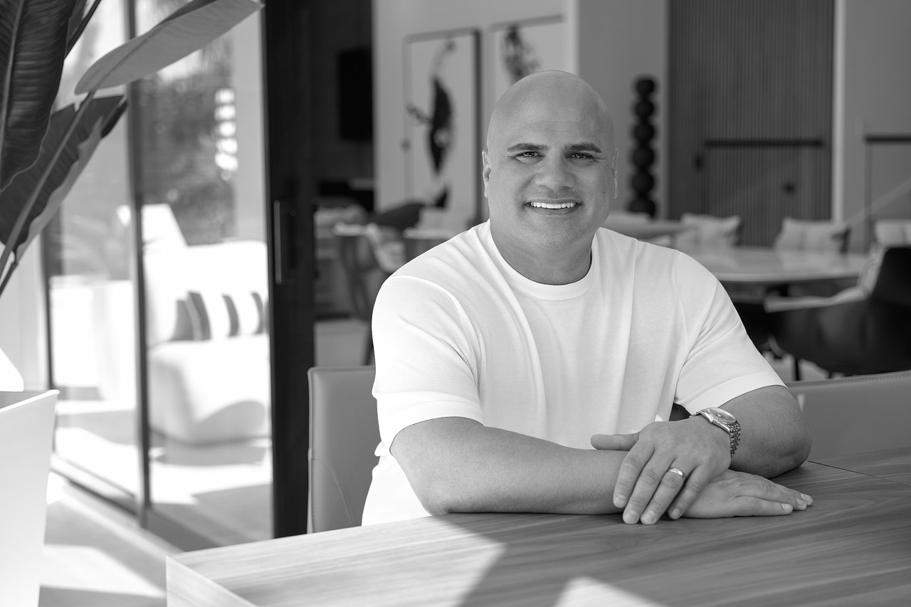 Marc Coriaty smiles in a white shirt, seated at a table in a modern interior.