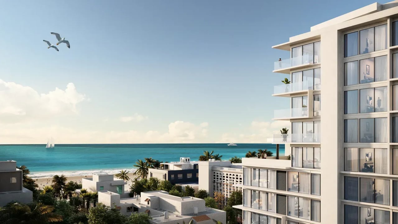 Modern white residential tower overlooking a turquoise beach, with glass balconies and palm-lined buildings below.