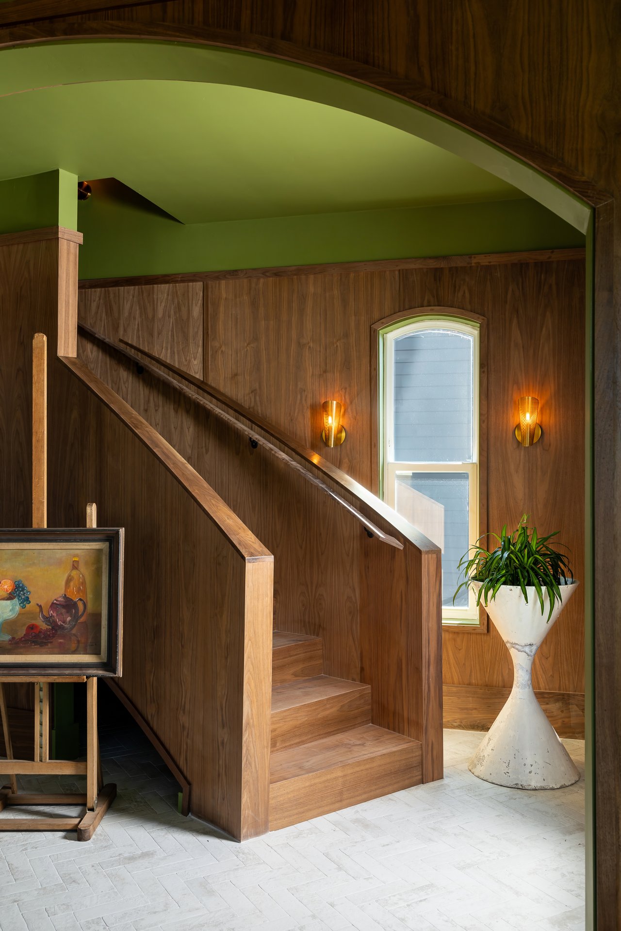 Wood-paneled foyer with a staircase, green arched ceiling, tall window, and glowing sconces.