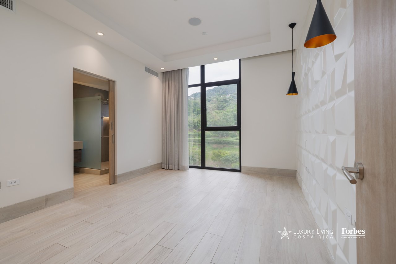 Vacant bedroom with a geometric accent wall, ensuite bathroom doorway, and a tall window overlooking green hills.