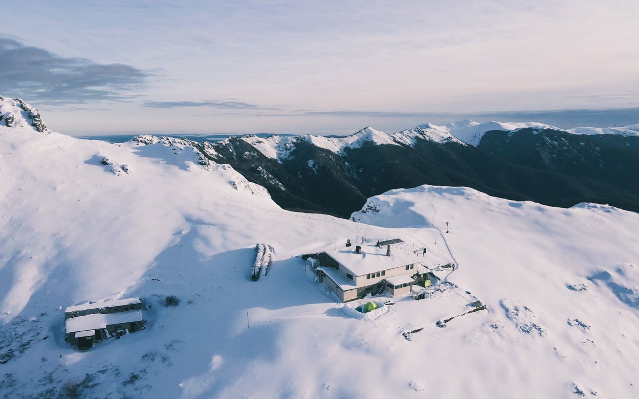 Mountain lodge and outbuilding surrounded by snow, with distant snow-covered peaks.