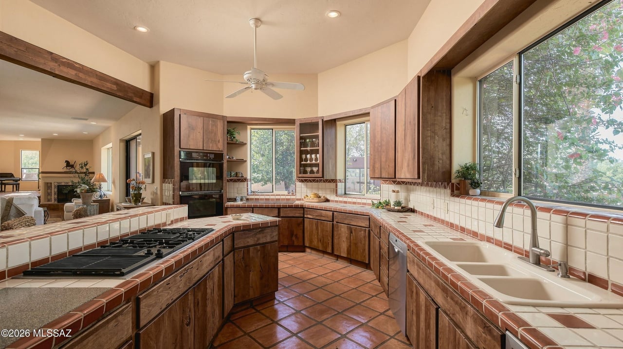 Kitchen with dark wood cabinets, tiled counters, gas cooktop, double ovens, and large windows overlooking trees.