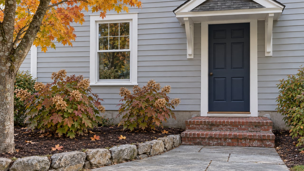 Gray clapboard home with a navy front door, paired entry brackets, brick steps and stone-edged autumn hydrangeas.