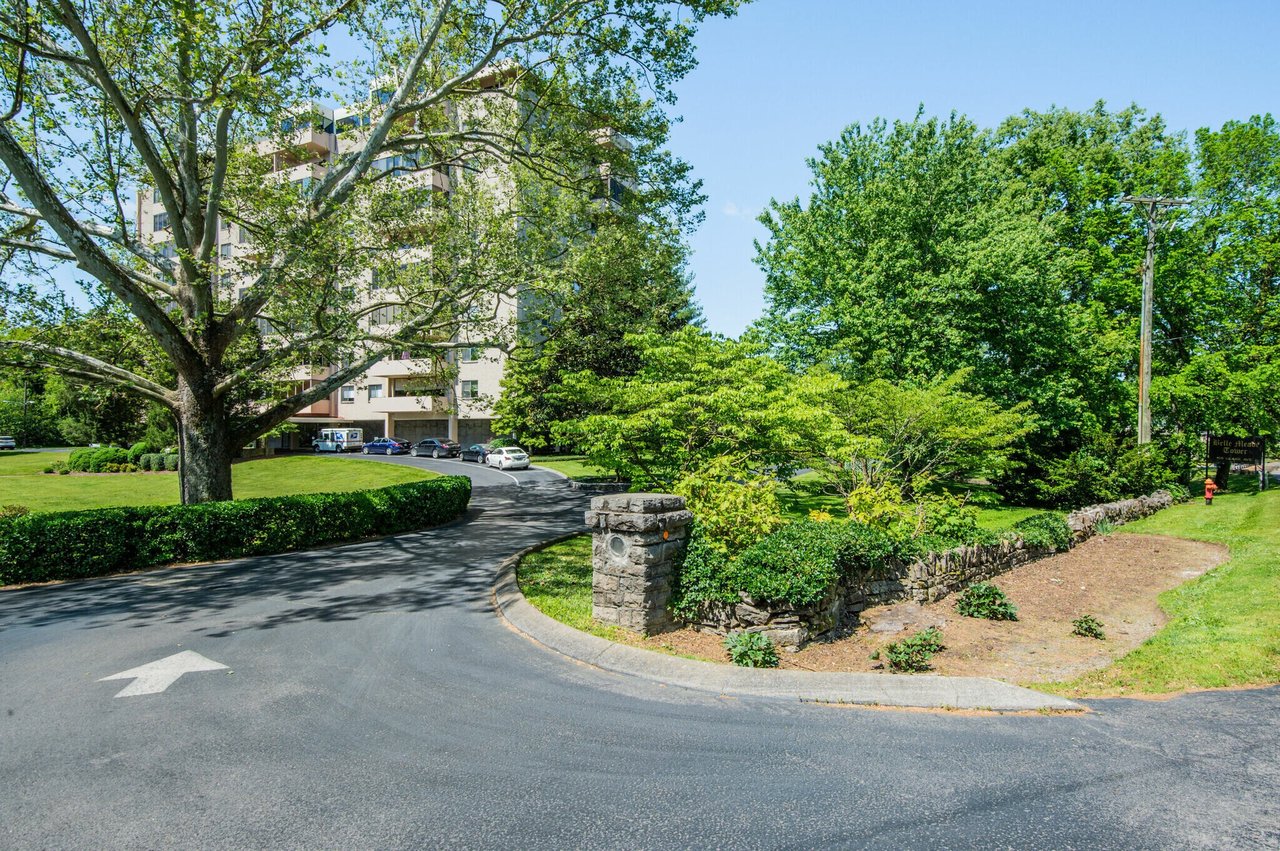 Residential building entrance with a curved driveway, stone marker, and mature trees.