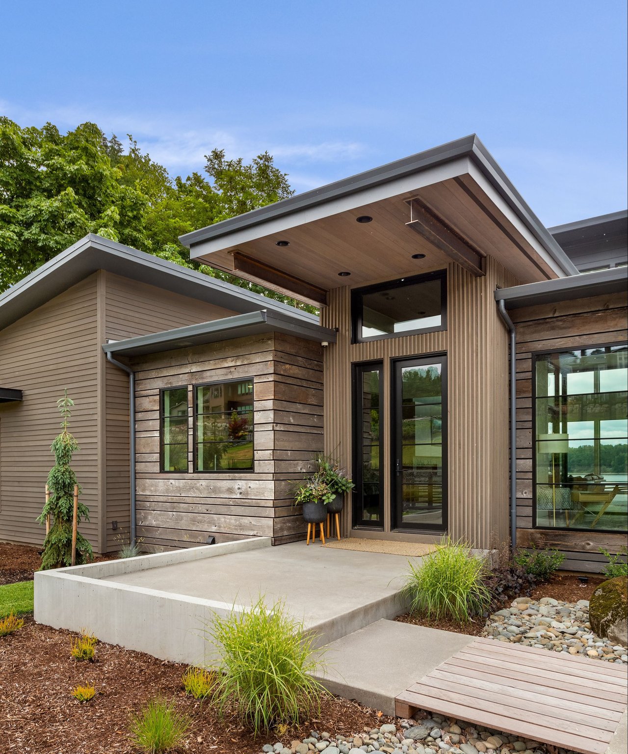 Modern wood-sided home with a broad concrete entry terrace and large windows overlooking water.