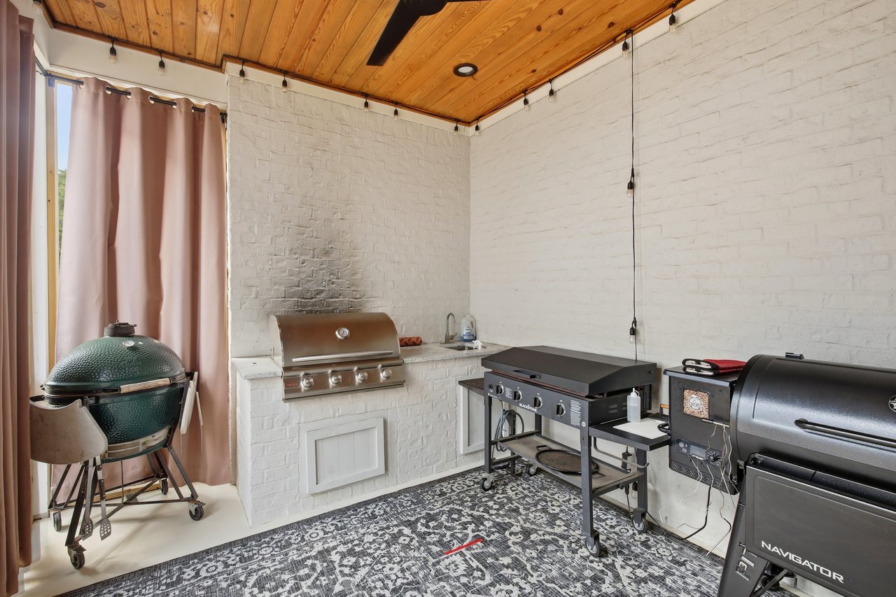 Covered outdoor kitchen with multiple grills, white brick walls, and a wood-plank ceiling.
