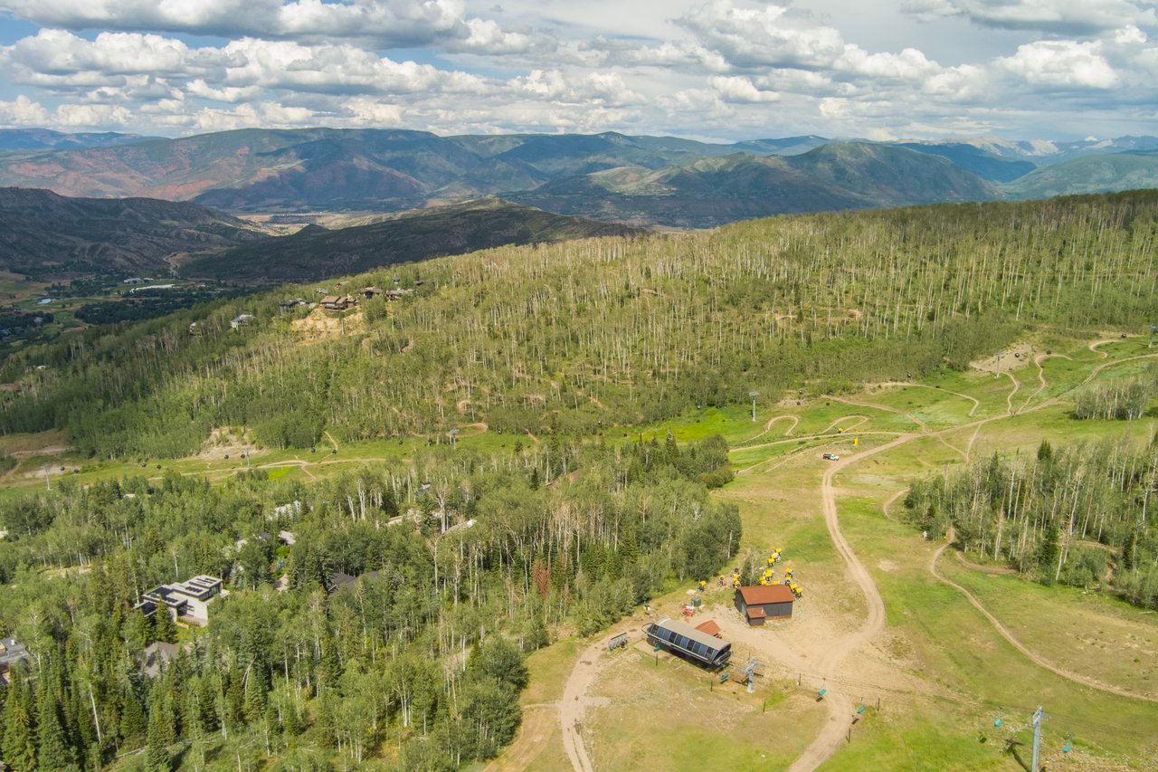 Aerial view of a wooded ski slope, with winding trails, lift infrastructure, and mountain ranges beyond.