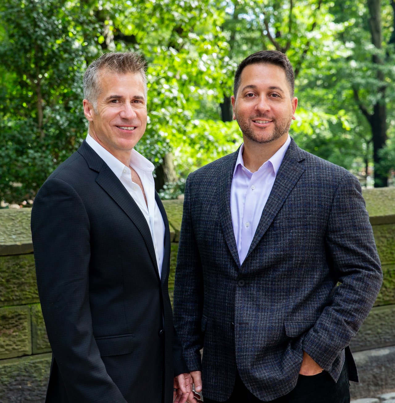 Two real estate agents in suits pose together outdoors against leafy greenery.