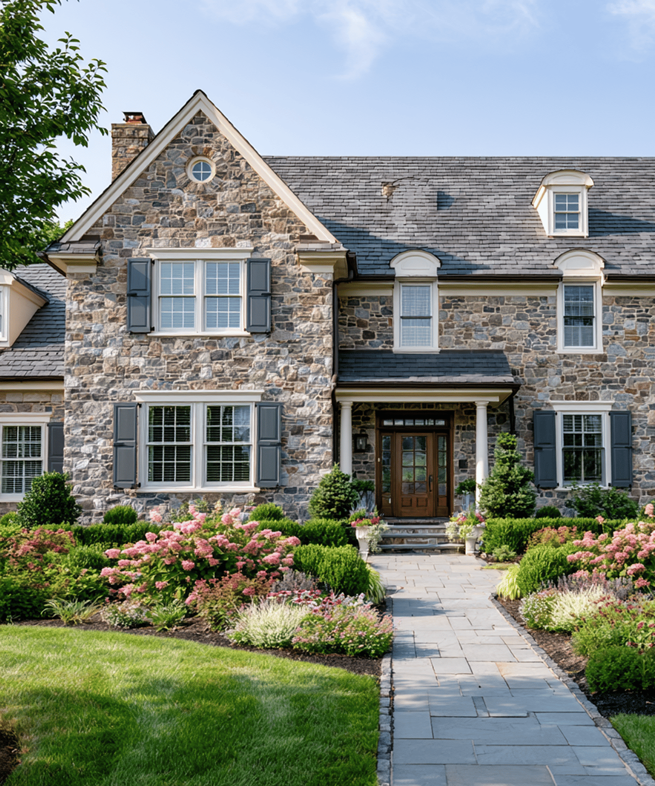 Stone house with gray shutters, a paved front path, and colorful landscaped gardens.