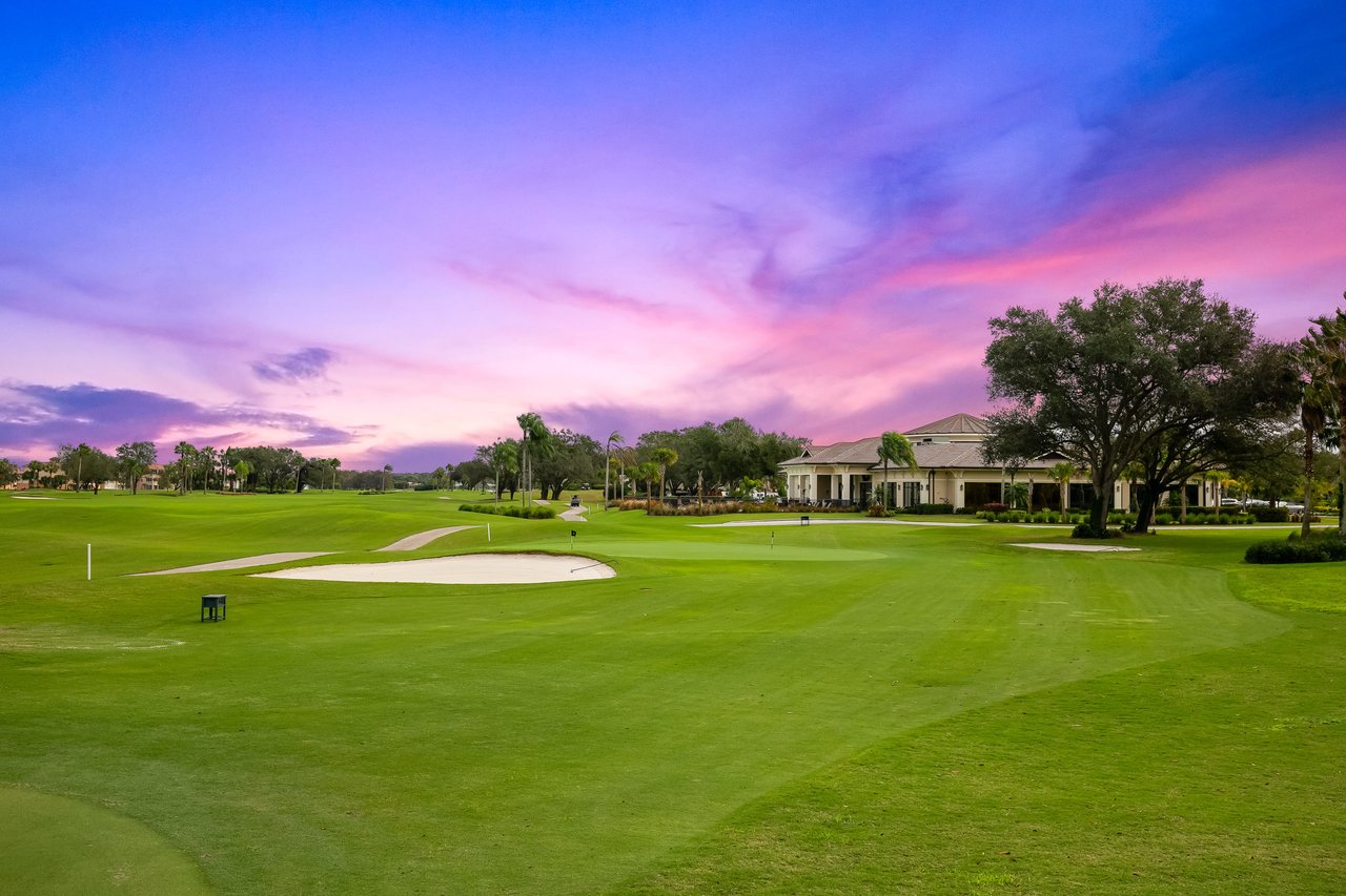 Golf course with a sand bunker and clubhouse beneath a vivid pink and purple dusk sky.