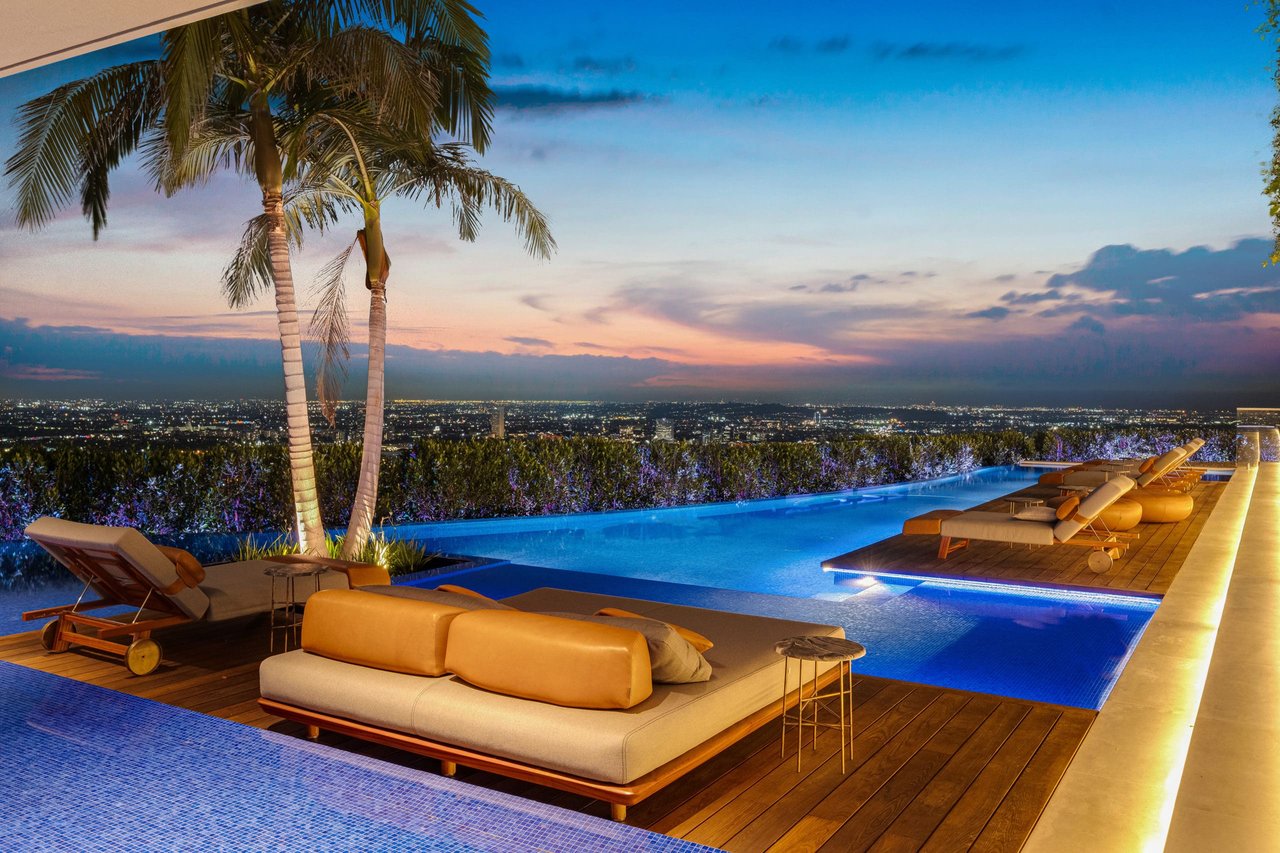 Infinity pool terrace with palm trees, loungers, and a city skyline at dusk.