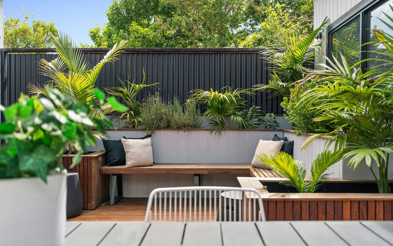 Outdoor patio with built-in wooden benches, pale cushions, and lush tropical planters.