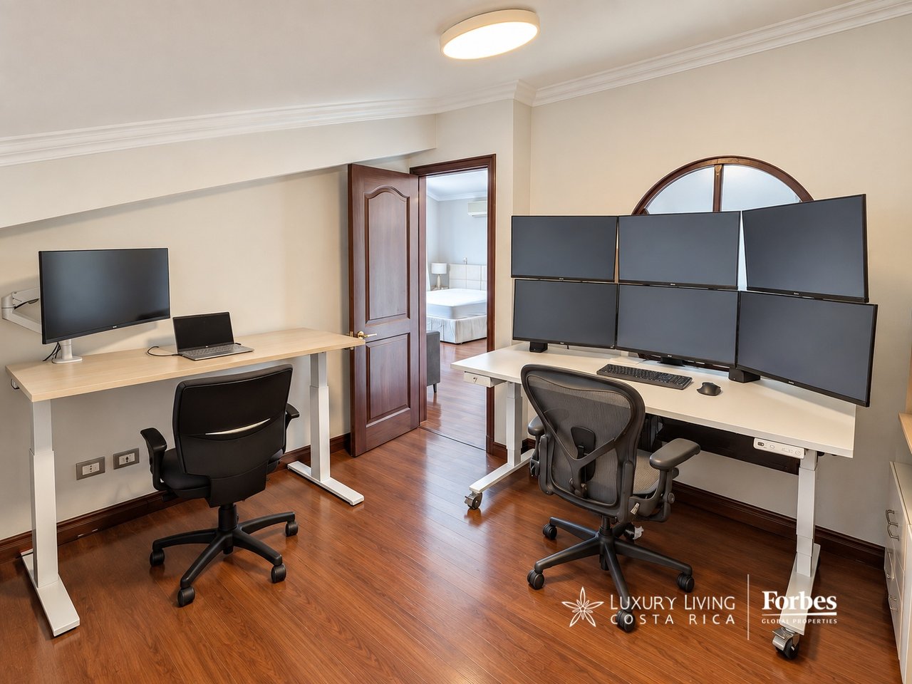Furnished home office with two desks, a six-monitor workstation, and an open doorway to a bedroom.