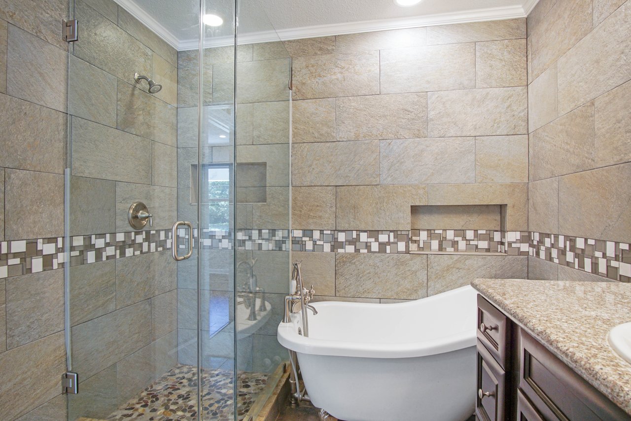 Modern bathroom with stone tile walls, a glass shower, and a white clawfoot bathtub. A decorative tile strip adds contrast. Warm, elegant ambiance.